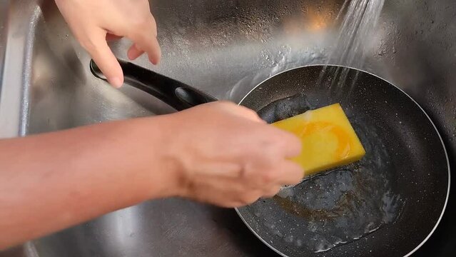 Unas manos de mujer lavando  un sart&eacute;n  con una esponja amarilla con jab&oacute;n l&iacute;quido y  agua en un fregadero