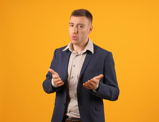 A man in a suit seems puzzled or questioning, hands spread out, against a yellow backdrop.
