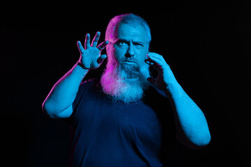 A man with neon pink and blue beard lighting, holding up glasses to his face, dark background, looking curious.