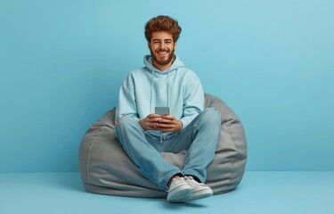 Against a blue backdrop, a full-size image of a perceptive and optimistic man wearing jeans and a sweater sits on a bean bag and looks at his smartphone.