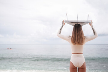 Banner back view of a young sexy woman standing on a beach near the ocean wearing a white surf bikini and holding a surfboard on her head. Happy sporty young female surfer. Let's go surfing