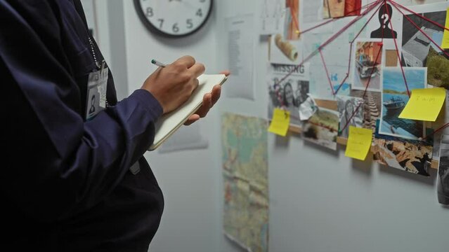 African american detective man taking notes in an office with an investigation board covered in evidence photos and maps.
