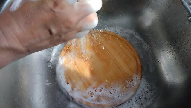 Unas manos de mujer lavando  una tabla de madera redonda con agua en un fregadero