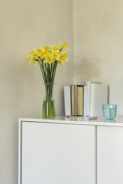 Vertical image of white dresser, blue glass of water, spring flowers in vase and stack of book. Flowers indoors. Fresh spring at home