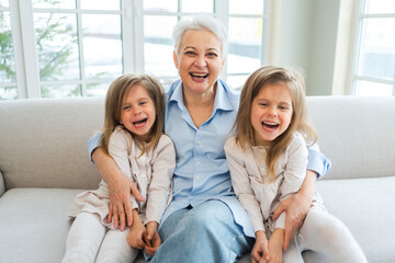 Fototapeta na wymiar Happy family at home. Two little girls sisters twins grandmother enjoying time together. Good time at home. Grandma granddaughters child kids emotional bonding hugging together. Family generations