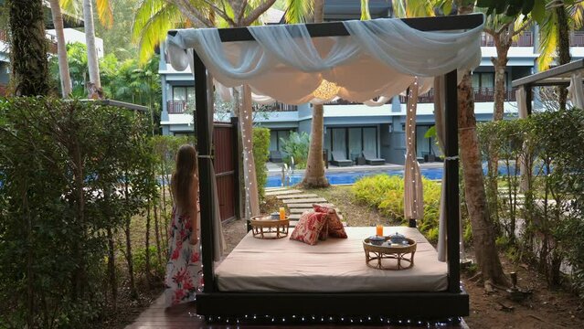 Elegant outdoor cabana set for romantic dinner surrounded by tropical plants at upscale resort, with woman in floral dress looking towards luxurious setting. Resort vacation and relaxation.
