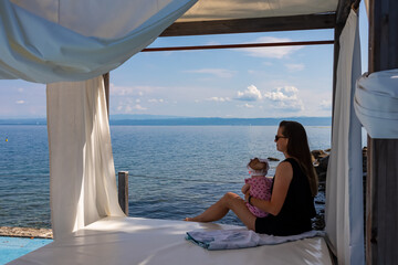 Woman holding a baby in lounger canopy beach bed in luxury hotel with scenic view of idyllic...