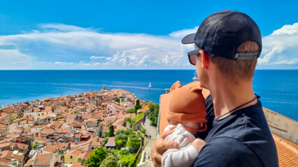 Fototapeta na wymiar Man holding baby enjoying scenic aerial view of coastal town Piran, Primorska, Slovenia. Shimmering azure waters of Adriatic Sea. Tranquil Mediterranean atmosphere. Exploring Slovenian Coast in Istria