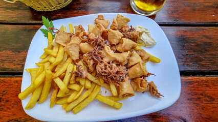 A plate of fried calamari served with a piece of lemon and garlic sauce. The dish is prepared on a white, round plate and located on a wooden table. Mediterranean cuisine. Frutti di mare.
