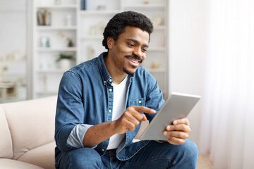 Smiling Black Man Using Digital Tablet On Couch in Bright Living Room
