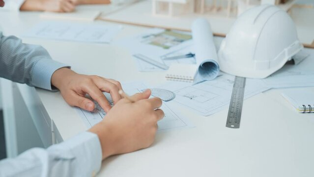 Close up of skilled architect engineer hand draws blueprint on meeting table with blueprint, architectural equipment, house model and safety helmet scatter around. Focus on hand. Immaculate.