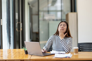 Obraz premium A young woman yawns while using a laptop at her desk. being tired during a busy work day
