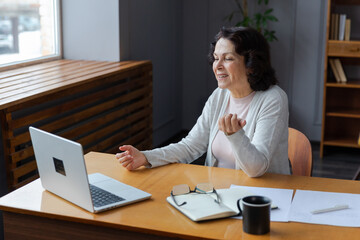 Happy middle aged senior woman sit with laptop talk on video call with friends family. Laughing mature old senior grandmother having fun talking speaking with grown up children online