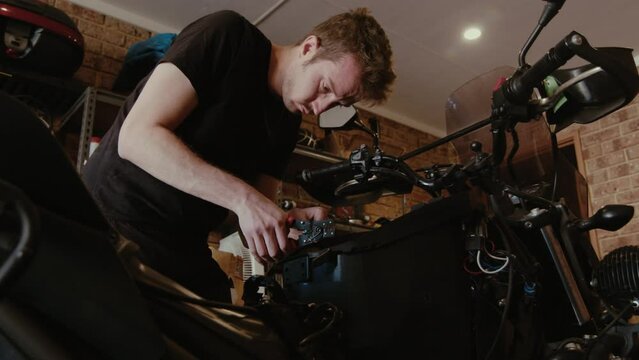 A focused young man adjusts the engine parts of a motorcycle, working with precision in a brick-walled workshop.