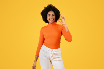 Contented woman with afro hair in orange turtleneck and white pants makes the 