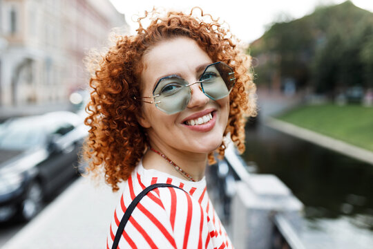 Back view outdoor portrait of positive happy female with bright curly hair in funny blue sunglasses turning her head by name to camera, strolling along city canal, admiring historical spots
