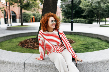 Happy cheerful millennial curly-haired redhead female sitting on stoned bench in city park, waiting for her friend to come after long time not seeing each other, enjoying fresh air and beautiful view - 779143696