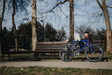 A focused businessman sits with his laptop on a park bench next to his bicycle, working remotely in...