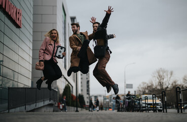 Three cheerful young business people jumping and celebrating outside an office building, showing...