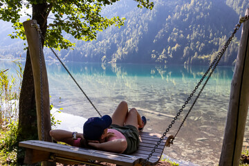 Woman relaxing on wooden swing with scenic view of alpine landscape at east bank of lake...