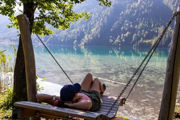 Fotobehang Ontspanning Woman relaxing on wooden swing with scenic view of alpine landscape at east bank of lake Weissensee, Carinthia, Austria. Tranquil forest in remote untouched nature in summer. Pristine turquoise water  © Chris