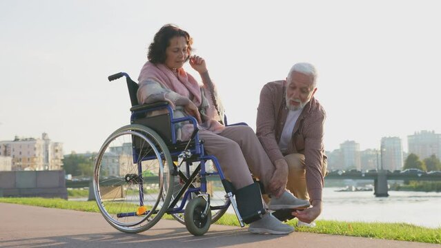 Senior woman in wheelchair walking with caregiver old man on road. Elderly family couple man supporting taking care of paralyzed woman in chair for people with disability outdoor. Rehabilitation