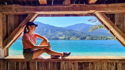 Woman sitting on edge of wooden platform looking at alpine landscape seen from east bank of lake Weissensee, Carinthia, Austria. Serene landscape in remote untouched nature in summer. Turquoise water - obrazy, fototapety, plakaty