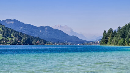 Obraz premium Scenic view of east bank of alpine lake Weissensee in Gailtal Alps, Carinthia, Austria. Bathing lake surrounded by mountain ranges of Austrian Alps. Serene landscape amidst untouched nature in summer