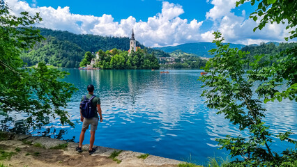 Tourist man with panoramic view of St Mary Church build on small island on alpine lake Bled, Upper Carniola, Slovenia. Serene landscape in Julian Alps in winter. Hills covered with lush green forest