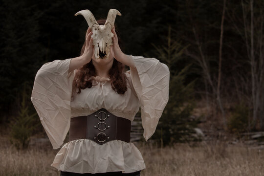 Beautiful Mysterious Ominous Witch Woman in Medieval Outfit holding Goat Skull over Face in Nature Forest with Space for Text