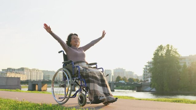 Happy senior woman in wheelchair raised hands enjoying freedom walking on road in city park outdoor. Old happy woman in chair for people with disability has a hope. Disability disease rehabilitation