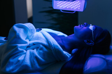 Young woman in glasses having blue LED light facial photodynamic therapy treatment in beauty salon. Female client having non-invasive type of phototherapy. Beauty, healthcare and wellness concept.