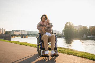 Old man in wheelchair walking with caregiver senior woman on road in park. Elderly family couple woman supporting embracing paralyzed man in chair for people with disability outdoor. Rehabilitation