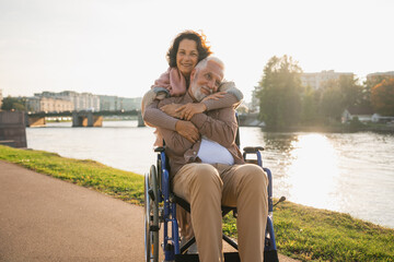 Old man in wheelchair walking with caregiver senior woman on road in park. Elderly family couple woman supporting embracing paralyzed man in chair for people with disability outdoor. Rehabilitation