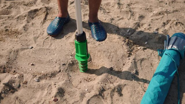 Caucasian man screwing screw base of beach umbrella into sand on ocean beach close up. Preparing place to relax on sea coast on hot sunny day and being aware of shade. 
