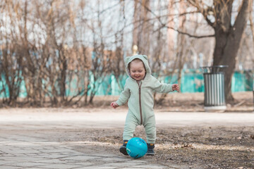 A handsome, cheerful little boy of two years old walks on the street in the spring. A kid in a jumpsuit is playing ball. The concept of parenthood, childhood and family.