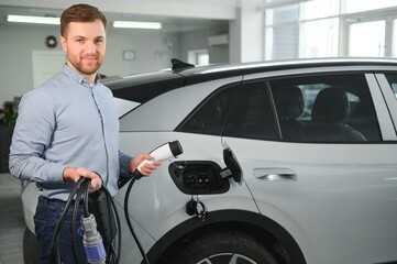 Handsome business man holding charging cable for electric car. Caucasian male stands near electric auto in dealership