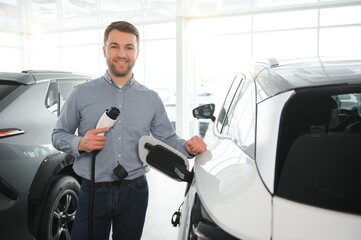 Beard man trying a new charging cable with a car charging station at the motor dealership. Concept of buying electric vehicle. Smart ecological living