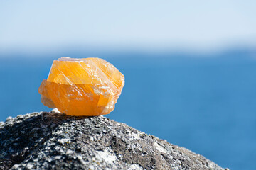 A close up image of an unpolished orange calcite crystal resting on a lichen covered rock with a blue ocean background. 