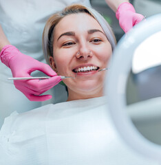 Portrait of smiling female gazing at mirror in stomatology chair and graceful dentist's palms in pink gloves with excavator and mirror medical tools. Health care and medicare industry concept image