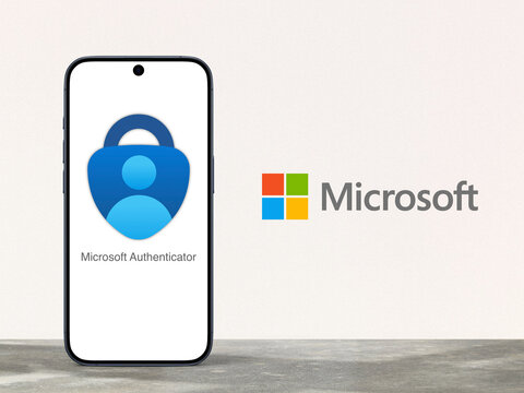 Microsoft Authenticator App logo is displayed on a modern smartphone standing on a desk, Microsoft logo next to it, fingerprint, face recognition, PIN, safety, security, prove account