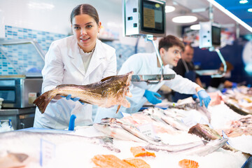Smiling salesgirl in work clothes holding in hands cod fish in fish shop - obrazy, fototapety, plakaty