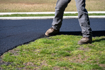 Closeup of a road construction worker using is foot packing down the new asphalt at the edge between with newly paved road and a lawn in someone’s yard
