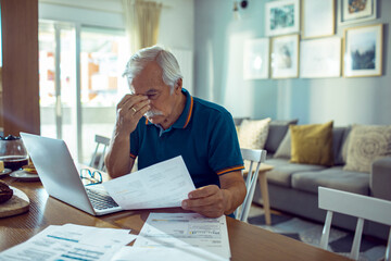 Senior man with glasses reviewing bills and using laptop at home