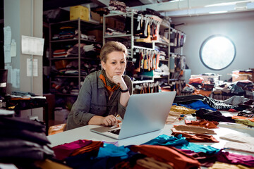Female fashion designer working on laptop in textile studio with fabric samples