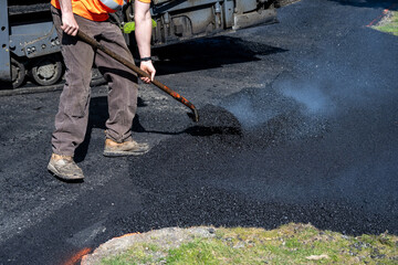 Road crew worker moving hot asphalt with a shovel at the intersection of a driveway and the street, road construction project
