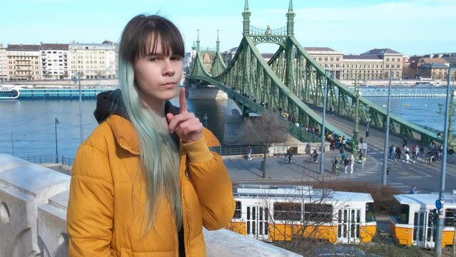 Girl point with finger near mouth. A view of young modern girl point her finger near mouth on the balcony against Budapest view. A concept of keep silence in the big city.