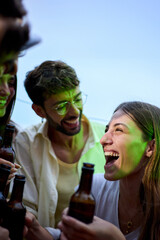 Vertical. Excited happy group friends toasting bottles of beer at dusk at rooftop party. Gathering...