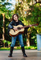 A beautiful little girl, in a rock style,in a leather jacket and jeans,stands on the street and plays an acoustic guitar
