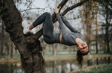 Athletic young female exercises by hanging upside down from a tree branch, showcasing strength and...